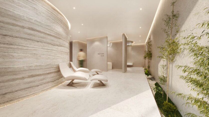 Salon de spa minimaliste au Marquis One Arjan avec des chaises blanches modernes, des murs texturés incurvés, des plantes d'intérieur, un éclairage doux et une silhouette floue passant devant le mur "SELF CARE CLUB". L'espace dégage une atmosphère calme et sereine.