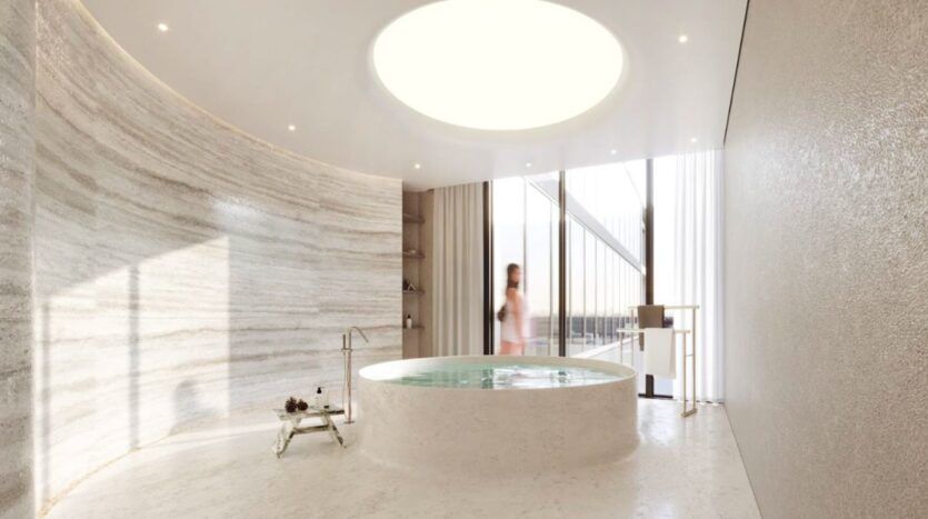 La salle de bains moderne et minimaliste du Marquis One Arjan présente une grande baignoire ronde au centre, un puits de lumière au-dessus, des murs en marbre incurvés et des fenêtres allant du sol au plafond. Une personne en tenue légère se tient près de la fenêtre.