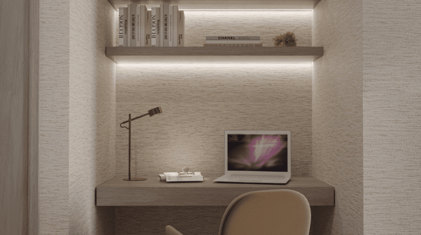 Un coin bureau minimaliste avec un bureau en bois, une chaise beige, un ordinateur portable ouvert, une lampe de bureau dorée, un carnet de notes et deux étagères avec des livres et une petite plante, le tout éclairé par un éclairage intégré doux.