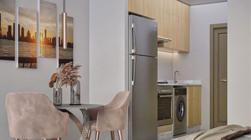 Une cuisine moderne avec des armoires en bois clair, des appareils électroménagers en acier inoxydable et une table à manger ronde avec deux chaises roses offre un style inspiré par Butterfly Towers Arjan. Un tableau mural représentant un coucher de soleil en ville et des fleurs séchées complètent l'ensemble.