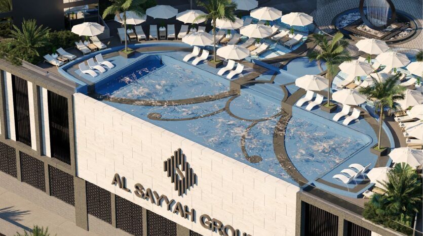 La piscine sur le toit du Butterfly Towers Arjan est agrémentée de chaises longues, de parasols, de palmiers et de l'inscription "AL SAYYAH GROUP" sur le mur extérieur de l'immeuble. Le design moderne comprend deux piscines reliées entre elles et de nombreux espaces de détente.