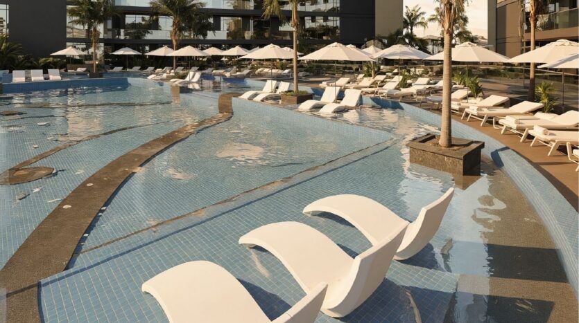 Une piscine avec des chaises longues blanches immergées, des palmiers environnants et de nombreux transats ombragés par de grands parasols blancs, avec en toile de fond l'architecture moderne des Butterfly Towers Arjan et d'élégants sièges au bord de la piscine.