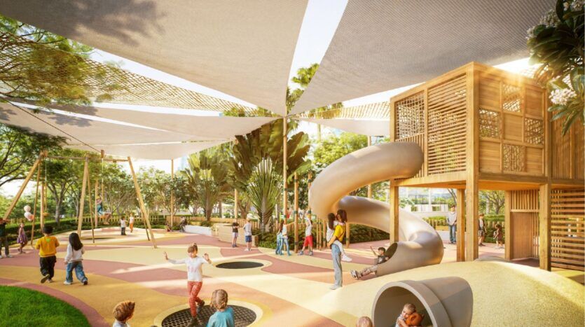Children play in Sierra by Iman's colorful outdoor playground, with slides, swings, climbing structures and shade awnings, all surrounded by greenery and trees on a sunny day.