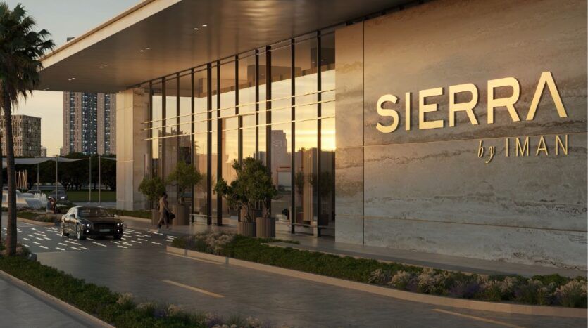 A modern building with a glass façade and a large "sierra by iman" sign stands amidst a lush green landscape, a car at the entrance and the city skyline lighting up at sunset.