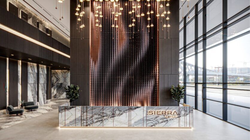 A modern office lobby with a marble reception counter inscribed "SIERRA by Iman", a dramatic wall with hanging lamps, large windows and tall plants on either side of the counter.