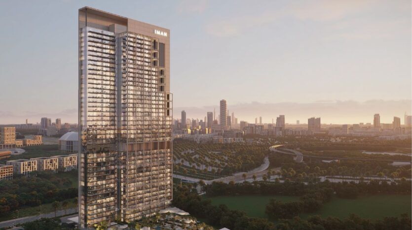 A large modern skyscraper bearing the inscription "EMAAR" rises above a verdant park, with the Sierra by Iman development and the city skyline in the background, against a hazy sunset sky.