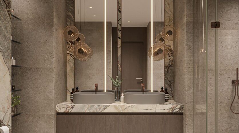 Modern bathroom with double sinks, marble countertop and large illuminated mirrors. Hanging lamps from the Sierra by Iman collection are suspended above the sinks, complementing the glass shower doors and sleek, contemporary design in neutral tones.