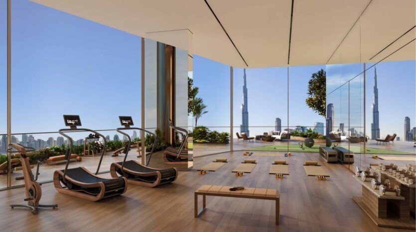 Salle de sport moderne avec sol en bois, tapis de course, tapis de yoga et chaises longues, dotée de grandes fenêtres allant du sol au plafond et offrant une vue panoramique sur l'horizon de la ville depuis la tour Sol Luxe, y compris l'emblématique Burj Khalifa.