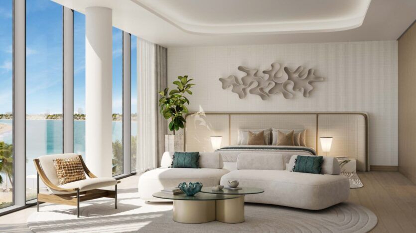 The modern, luxurious Palm Central room features floor-to-ceiling windows overlooking the beach. It features a curved white sofa, a bed with neutral linens, a glass coffee table, stunning wall art and an elegant potted plant.