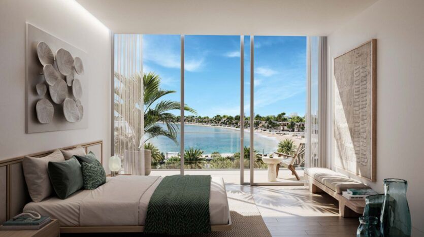 A modern room with a king-size bed, green accents and minimal decor - the best of Palm Central - features floor-to-ceiling windows offering panoramic views of a sunny beach, swaying palms and blue ocean water.