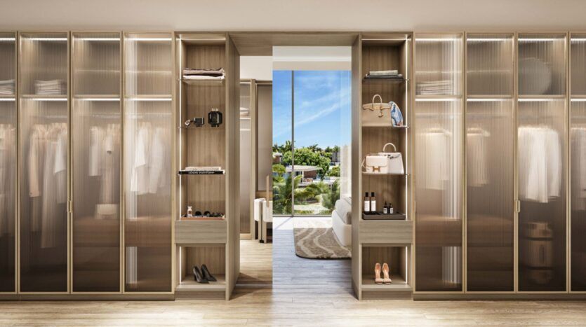 A modern Palm Central dressing room with glass closets, tidy clothes, shoes and accessories. An open door reveals a bright bedroom and a view of the greenery and blue sky outside.