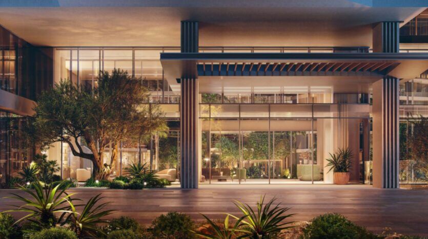 The modern entrance to the Palm Central building features large bay windows, lush greenery and plants. Warm interior lighting creates a welcoming atmosphere, complemented by outdoor landscaping featuring a variety of shrubs and small trees.