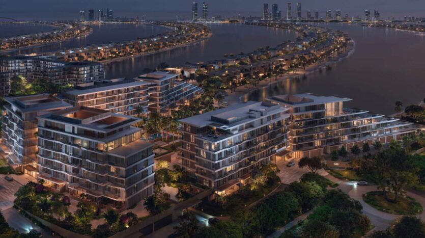 Nighttime aerial view of modern multi-story apartment buildings on Palm Central's waterfront, with illuminated windows, lush landscaping and palm trees, on a palm-shaped island, with the skyline and water in the background.