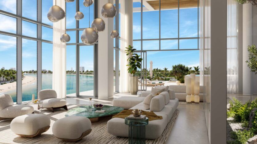 The luxurious, modern living room features floor-to-ceiling windows overlooking the beach and Palm Central's iconic palm trees. White sofas, elegant decor and large hanging lamps create an airy, elegant atmosphere bathed in natural light.
