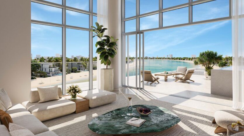 The Palm Central's modern living room, with large floor-to-ceiling windows, opens onto a sunny terrace overlooking a tranquil beach and blue waters. The room features a green marble coffee table, beige sofas and indoor plants.