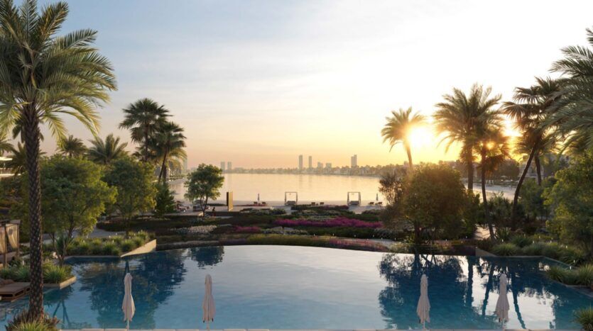 The Palm Central's serene pool, surrounded by lush gardens and palm trees, overlooks a calm waterfront at sunset, with the city's buildings visible across the water under a clear sky.