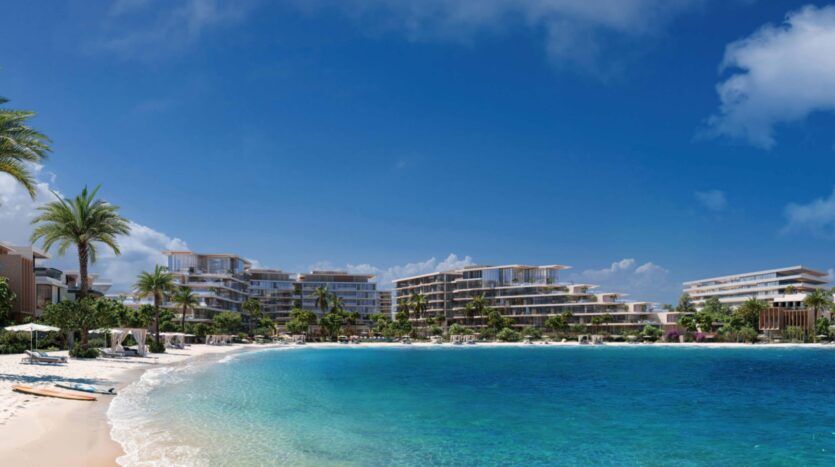 The modern Palm Central resorts, with their palm trees, line a sandy shoreline along clear blue water under a brilliant blue sky; sun loungers and parasols are set up along the water's edge.
