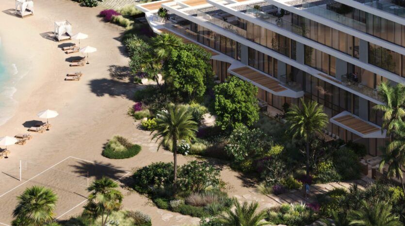 The Palm Central is a modern beachfront building with glass balconies overlooking a sandy beach lined with palm trees, lush vegetation and sun loungers shaded by white parasols near calm blue water.