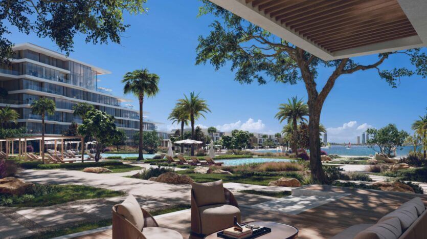 A luxurious outdoor lounge with modern chairs overlooks a pool, palm trees and landscaped gardens at Palm Central. Contemporary buildings and a waterfront are visible under a clear blue sky in the background.