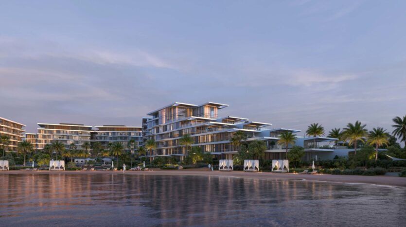 Palm Central, a modern beachfront resort, features several glass-walled buildings, palm trees and white cabanas along the shoreline, all beautifully reflected in the calm water at sunset.