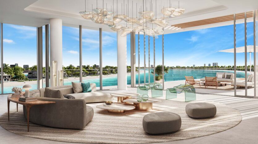 Modern, spacious Palm Central lounge with large windows overlooking a blue waterfront. Elegant furnishings include sofas, armchairs and a round rug. An outdoor patio with sun loungers and parasol is visible under sunny skies.