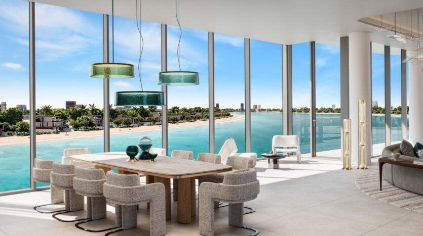 Modern dining room with large windows overlooking turquoise waterfront, sandy beach and iconic Palm Central views. The room features a wooden table, upholstered chairs, decorative lamps and contemporary decor.