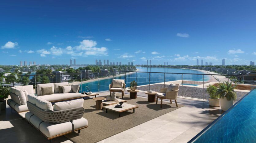 The Palm Central's luxurious outdoor terrace, with its cushioned seating, low tables and potted plants, overlooks a serene pool and vast waterside beach, under a clear blue sky. Modern buildings and lush greenery enhance the tranquil setting.
