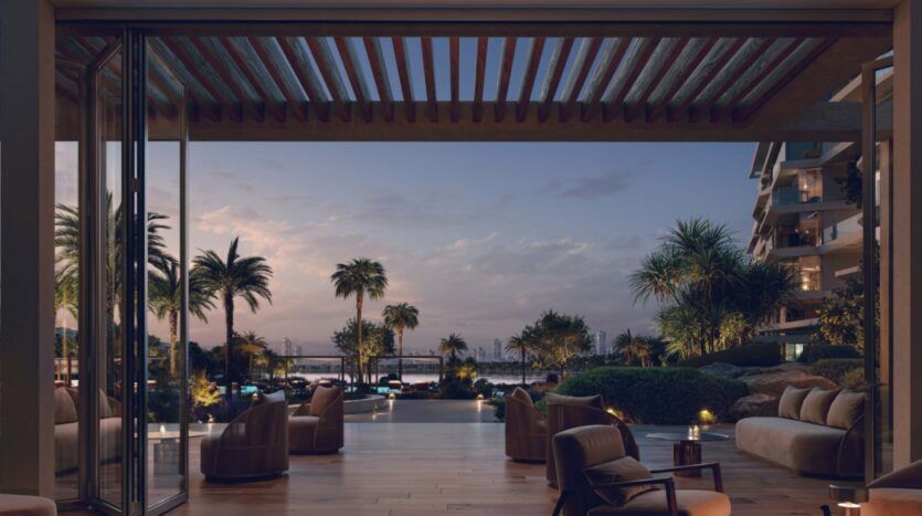 The Palm Central's open-air lounge, with modern seating and wooden flooring, offers views of the gardens, palm trees and waterfront at sunset. Warm lighting creates a relaxed, welcoming atmosphere, with views of the city skyline in the distance.