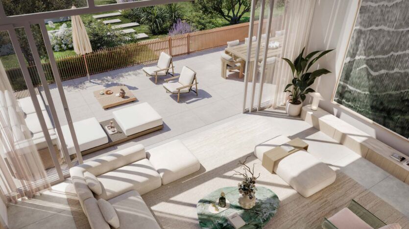 The Palm Central's bright, modern living room, with its white furnishings, opens onto a spacious patio adorned with lounge chairs, a dining table and greenery. Large windows let in natural light, creating an airy, welcoming atmosphere.