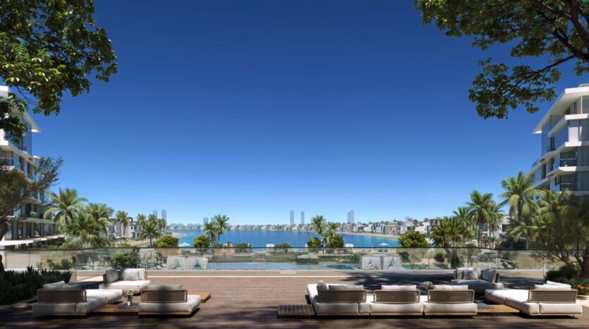 The Palm Central's luxurious terrace, complete with cushioned seating, overlooks a pool, palm trees and a bay dotted with urban buildings. Trees frame the relaxing tropical atmosphere under a clear blue sky.