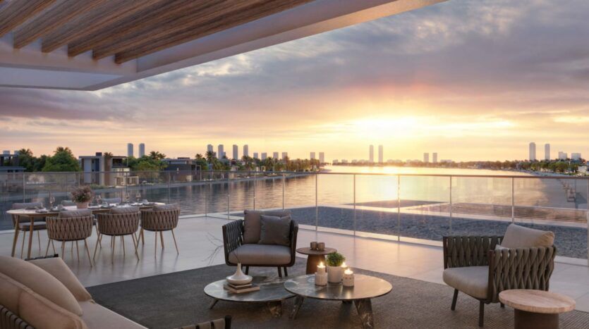 A modern terrace with elegant outdoor furniture overlooks a calm waterfront at sunset. The city skyline and Palm Central's iconic palm trees are visible in the distance, creating a tranquil, luxurious atmosphere.