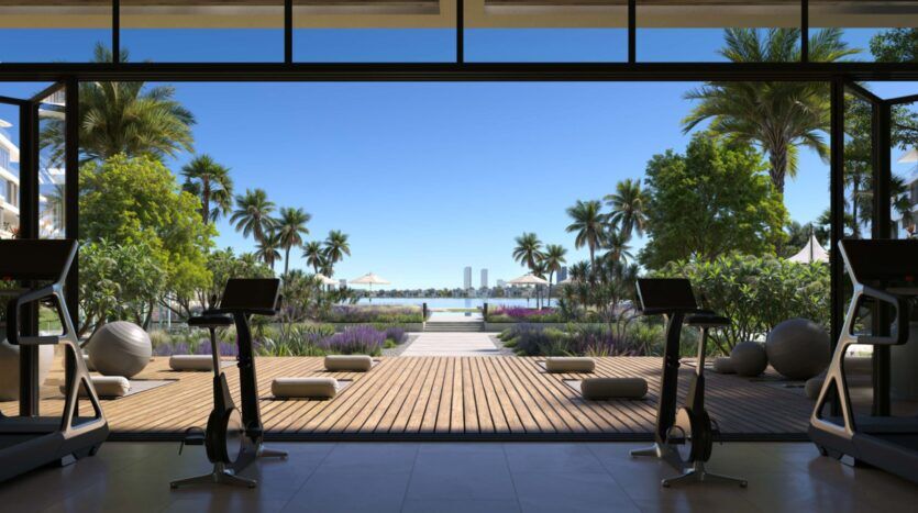 Open gym at Palm Central with exercise bikes facing large glass doors, overlooking a terrace, lush palms, gardens and bright blue skies with modern buildings in the distance.