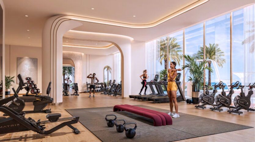 Une salle de sport moderne au Breez by Danube Maritime City avec de grandes fenêtres donnant sur les palmiers et l'océan, avec des vélos d'exercice, des tapis roulants, des poids, et plusieurs personnes s'entraînant dans un environnement clair et spacieux avec de la lumière naturelle.