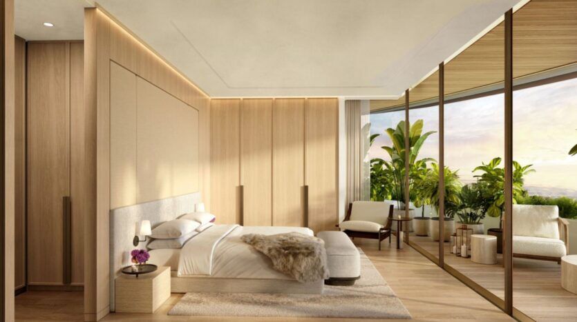 La chambre à coucher moderne de l'hôtel The Edition est dotée de murs et de sols en bois clair, d'un grand lit, de tables de chevet, d'un fauteuil confortable et de fenêtres allant du sol au plafond qui s'ouvrent sur un balcon luxuriant agrémenté de plantes vertes et de sièges d'extérieur. La lumière naturelle remplit la pièce.