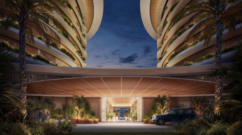 L'entrée d'un hôtel de luxe moderne au crépuscule, encadrée par des balcons incurvés avec de la verdure luxuriante et des palmiers, s'illumine d'une lumière chaude à côté d'une voiture garée. Le design ouvert de l'Edition offre une vue imprenable sur l'océan.