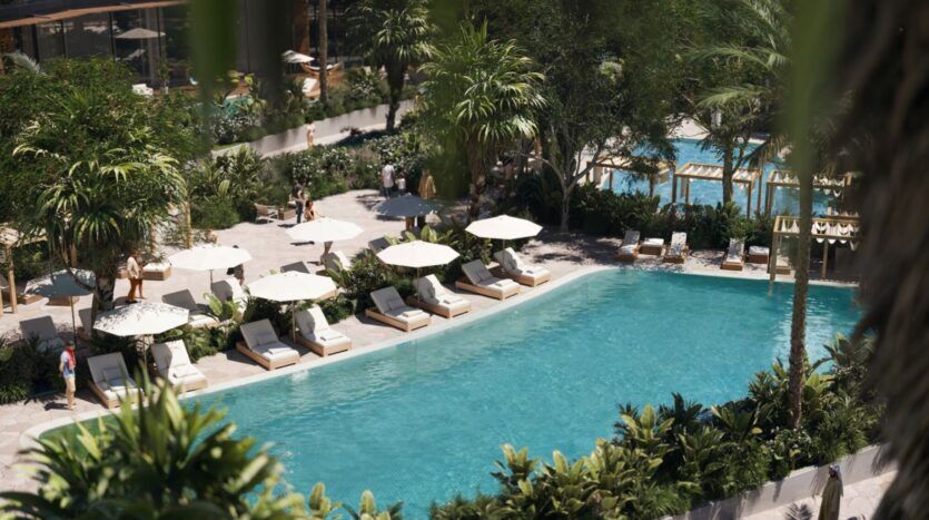 Une piscine extérieure luxuriante à The Edition, entourée de plantes tropicales, avec des chaises longues et des parasols blancs au bord de la piscine. Les gens se détendent et se promènent par un temps clair et ensoleillé.
