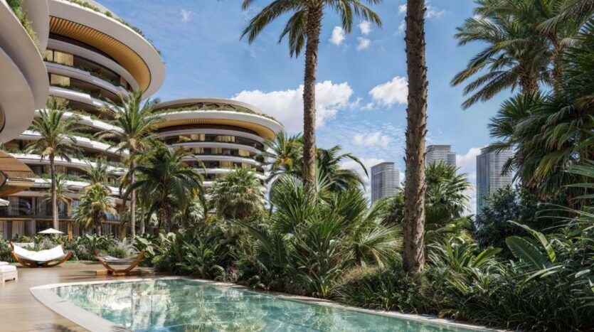 Des immeubles de luxe, dont The Edition avec ses balcons incurvés, s'élèvent derrière des jardins tropicaux luxuriants et des palmiers. Une piscine transparente scintille au premier plan, tandis que les gratte-ciel de la ville moderne se dressent sous un ciel d'un bleu éclatant.