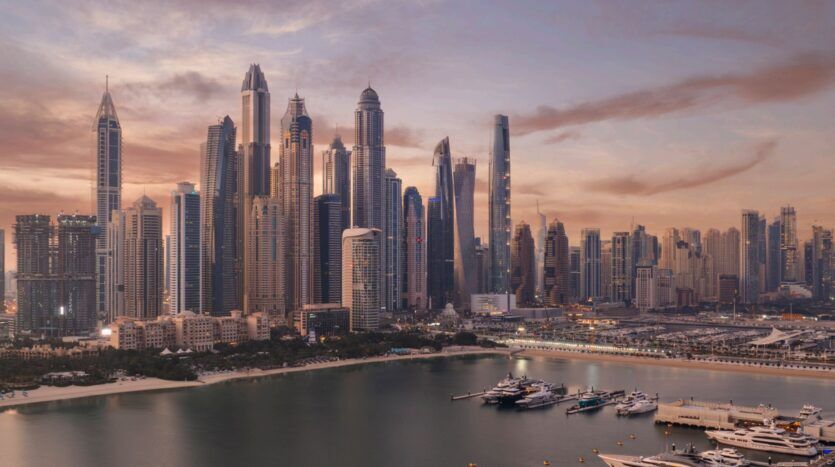 Une vue panoramique de la ligne d'horizon d'une ville moderne au coucher du soleil, avec The Edition au milieu de grands gratte-ciel, surplombe un port de plaisance rempli de yachts et d'eau calme, le tout sous un ciel rose et orange doux.