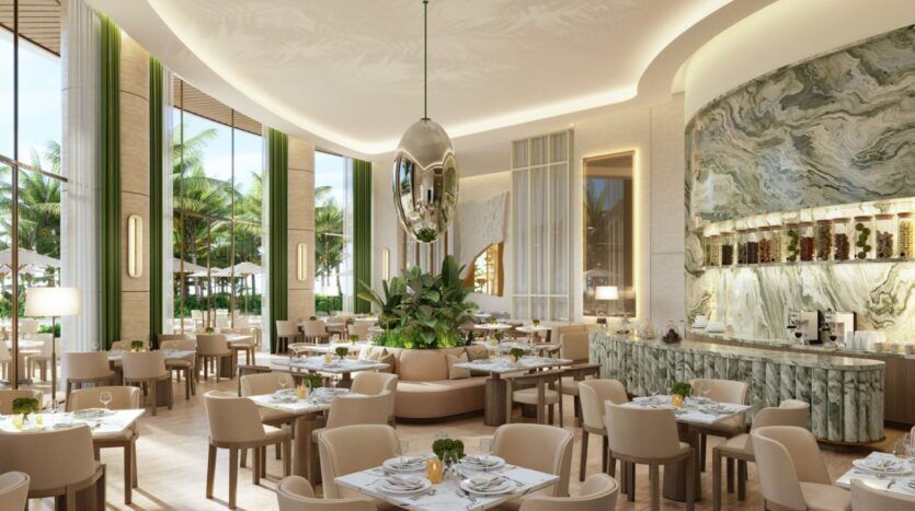 Ce restaurant moderne et élégant de l'Edition est doté de chaises et de tables beiges, d'un arrangement végétal central, de grandes fenêtres donnant sur les palmiers à l'extérieur, et d'un mur d'accent en marbre comportant des étagères et une station de café. Un éclairage doux crée une ambiance accueillante.