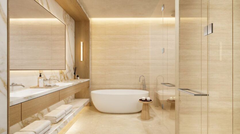 La salle de bains moderne de l'Edition présente des murs en pierre beige, une baignoire blanche indépendante, un tabouret en bois avec des bougies, un double lavabo avec un grand miroir et des serviettes soigneusement empilées sur des étagères ouvertes sous le comptoir.