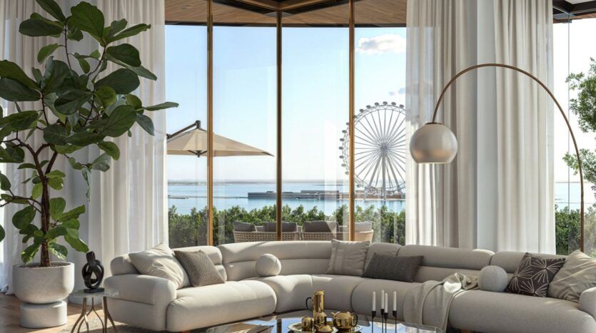 Le salon moderne de l'Edition est composé d'un grand canapé beige incurvé, d'une table basse en verre, de plantes luxuriantes et de hautes fenêtres offrant une vue sur la grande roue, l'océan et un patio avec un parasol. La lumière envahit cet espace aéré et élégant.