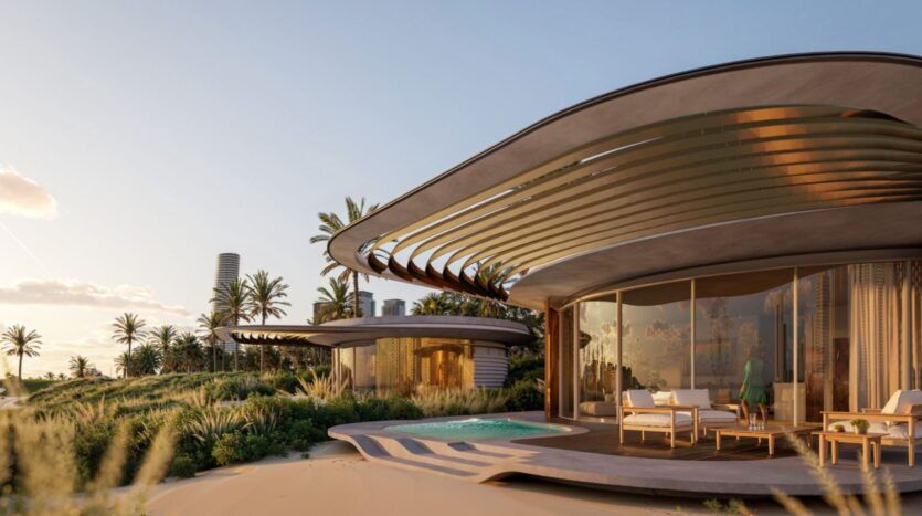 Une villa moderne en bord de mer à The Edition avec une architecture incurvée, de grandes baies vitrées, une piscine et des sièges extérieurs entourés de palmiers et de verdure sous un ciel dégagé. Une autre villa similaire est visible à l'arrière-plan.