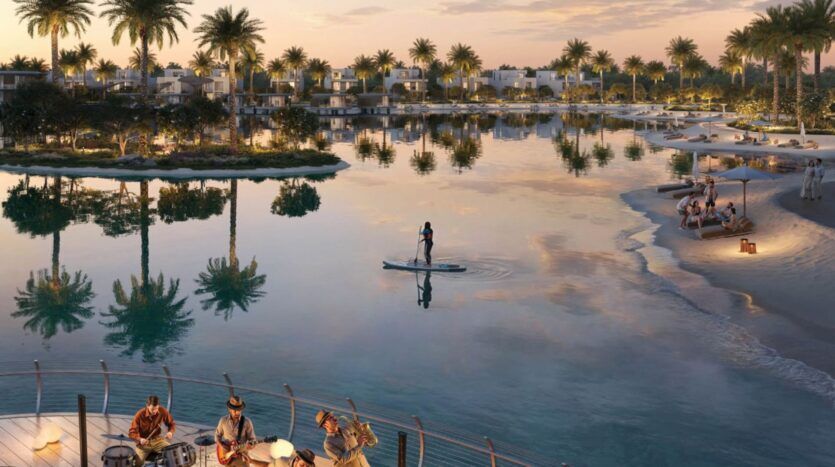 Une scène sereine au bord de l'eau au coucher du soleil à The Heights, avec une personne faisant du paddleboard sur une eau calme, des gens se relaxant sur une plage de sable, et des musiciens jouant sur une terrasse en bois entourée de palmiers et de villas modernes.