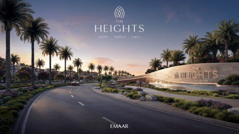 Une large route incurvée bordée de palmiers mène à l'élégant panneau d'entrée de The Heights, signé Emaar. Au coucher du soleil, on aperçoit en arrière-plan la verdure, les lumières et les bâtiments haut de gamme.