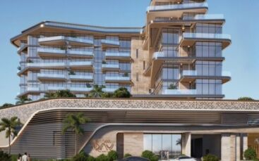 Un bâtiment moderne de plusieurs étages avec des balcons en verre incurvés, des murs texturés de couleur beige et des terrasses verdoyantes vous accueille à Flora Bay. Des palmiers et des voitures garées bordent l'entrée sous un ciel bleu clair.