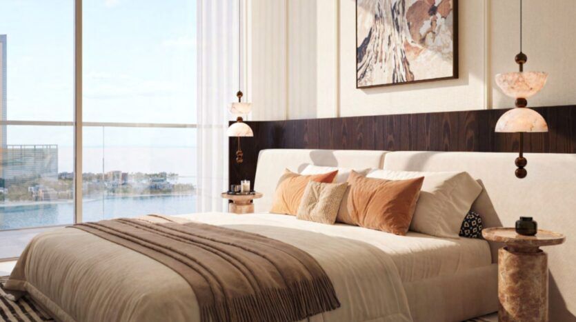 A modern bedroom in the Château with a large bed, brown and beige cushions, and a blanket. Hanging lamps, abstract wall art, side tables, and floor-to-ceiling windows offer views of the water and buildings outside.