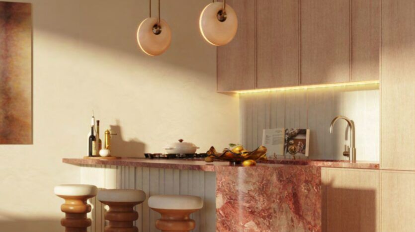 A modern kitchen with a pink marble countertop, three upholstered stools, pendant lights, and a Le Château-inspired backsplash with under-cabinet lighting. Decorative objects and kitchen items are displayed on the countertop.