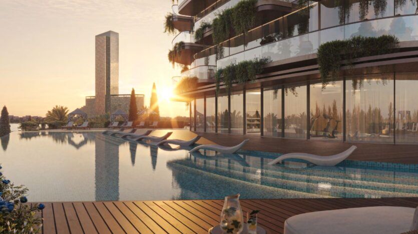 The Château's luxurious pool is equipped with modern sun loungers, a wooden deck, and a glass building at sunset. Greenery adorns the balconies, with a large skyscraper visible in the background.