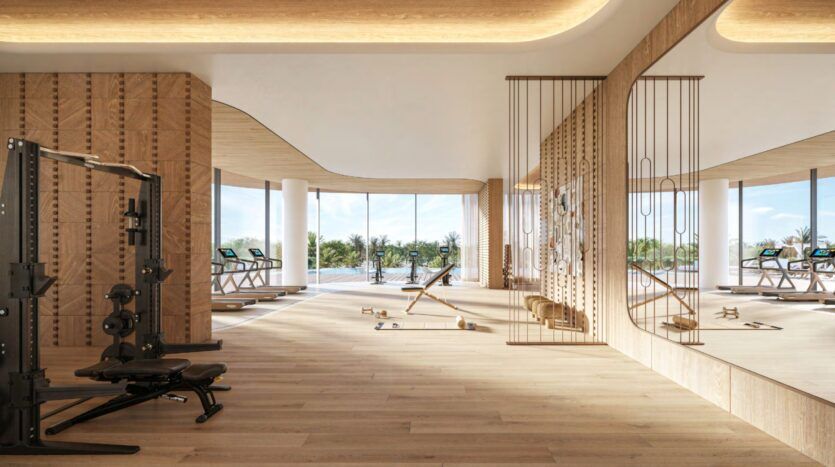 Modern gym at the Château with large windows, wooden floors, and training equipment including weight machines, benches, treadmills, and free weights. The space is bathed in natural light and overlooks palm trees and a swimming pool.