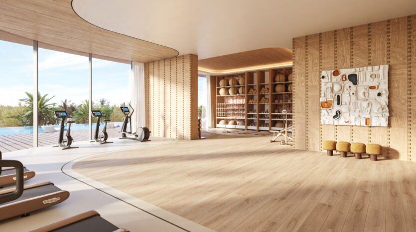A spacious and modern gym at the Château with wooden floors, large windows overlooking a swimming pool and palm trees, exercise machines, shelves with towels, and textured wall art. The room is bathed in warm natural light.
